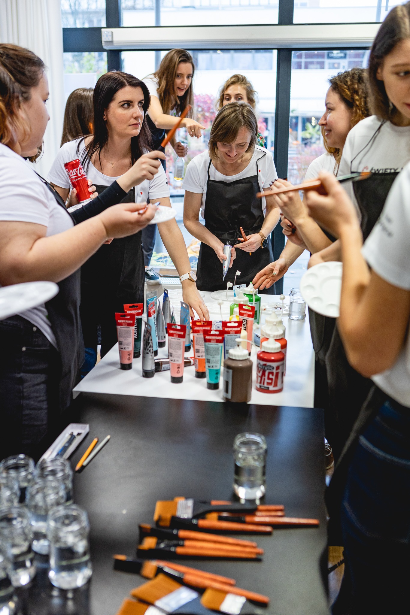 Grupa kobiet w fartuchach malarskich uczestniczy w warsztatach artystycznych, używając farb akrylowych i pędzli przy stole.