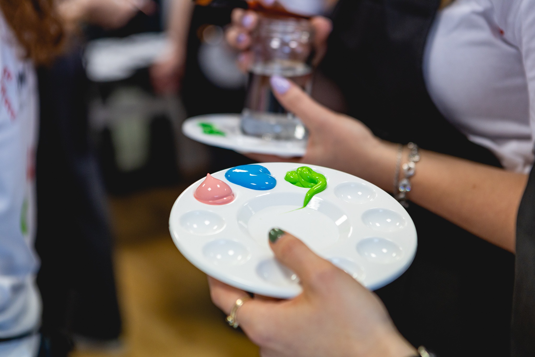 Paleta malarska z różową, niebieską i zieloną farbą, trzymana przez osobę z fioletowym lakierem na paznokciach, w tle szklanka z wodą.