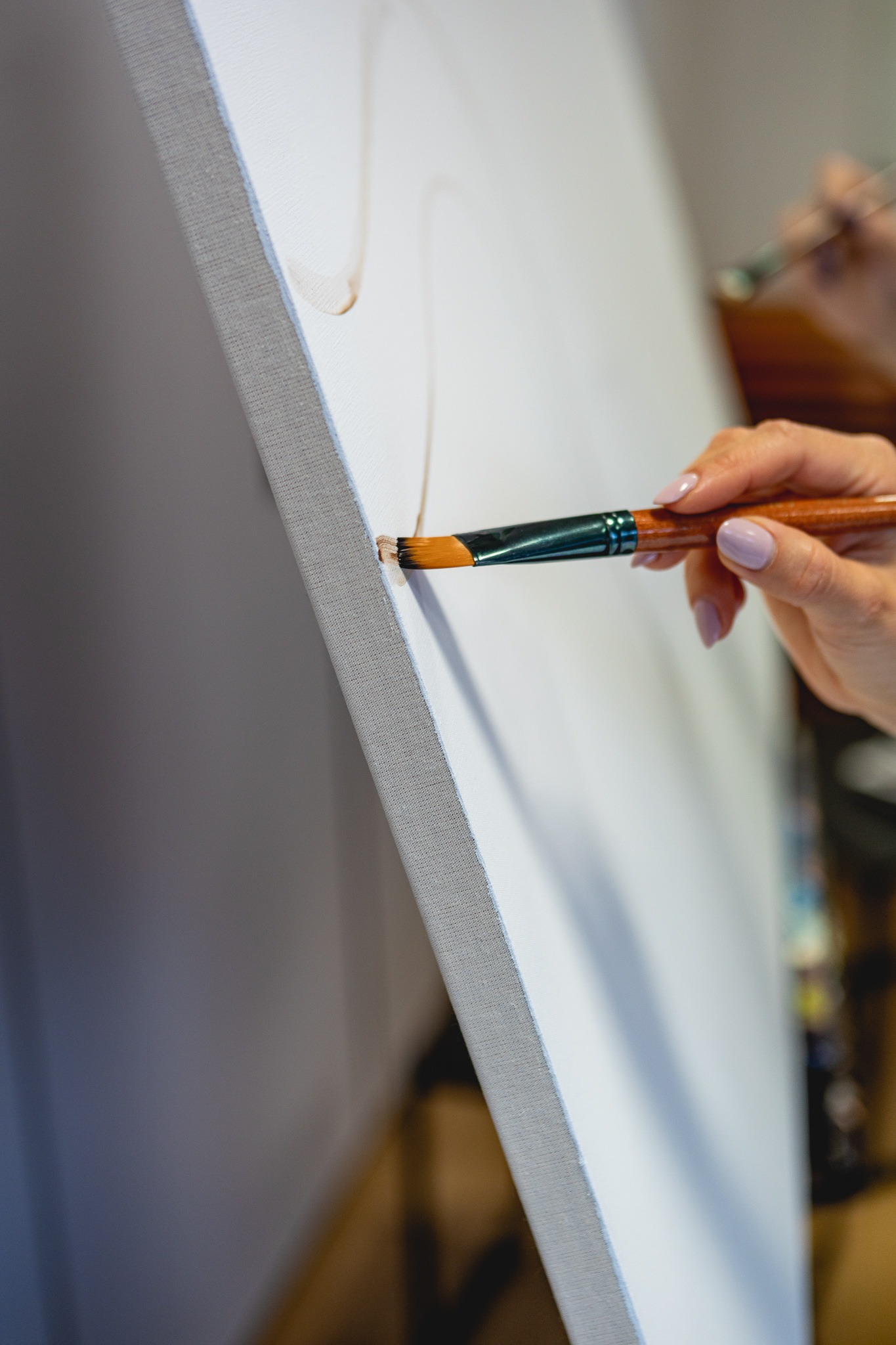 Artysta maluje pędzlem brązową linię na białym płótnie naciągniętym na ramę, widoczny fragment dłoni z jasnoróżowym manicure.