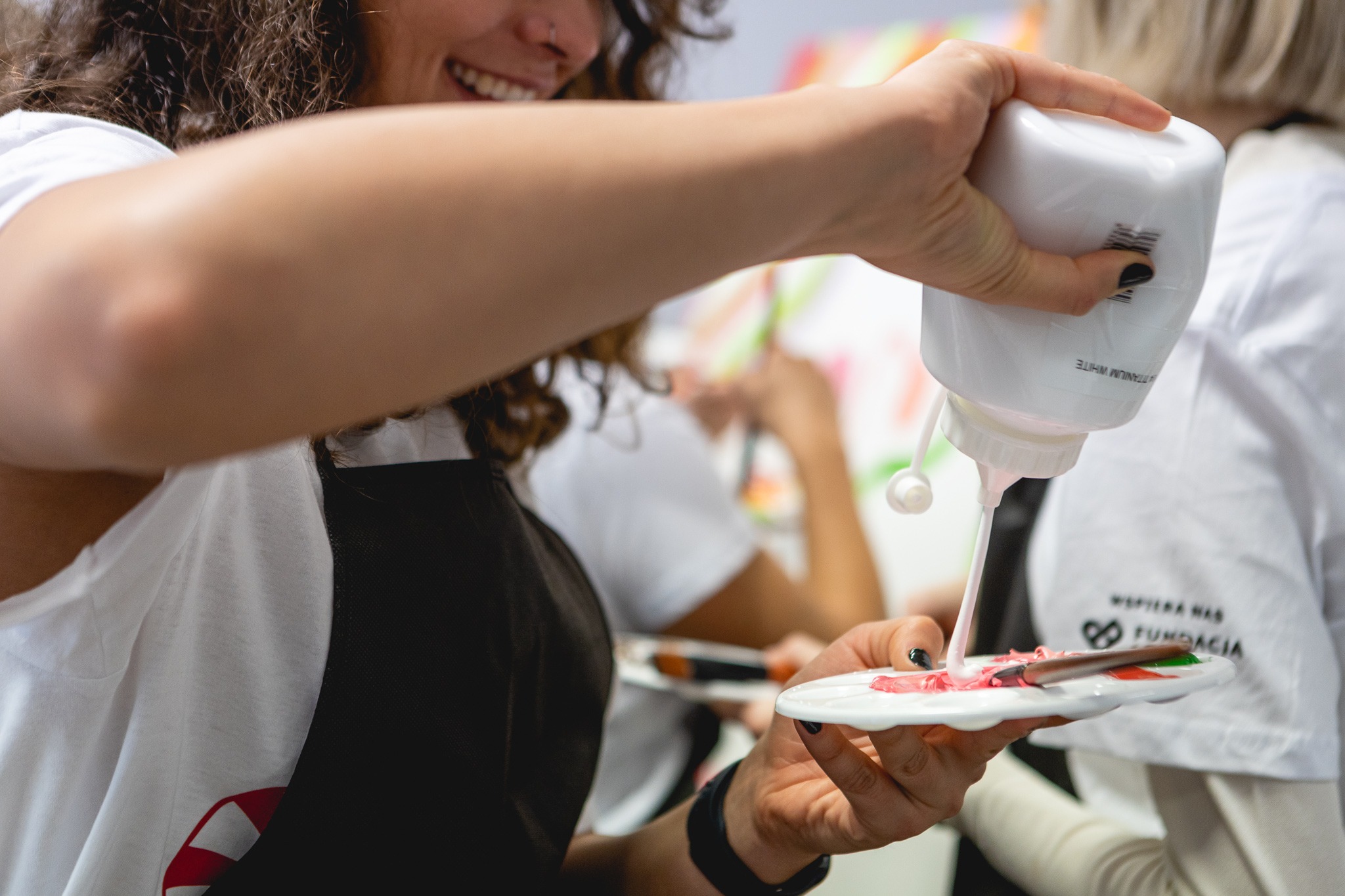 Uśmiechnięta kobieta w fartuchu wylewa białą farbę z tuby na paletę z mieszanką kolorów, prawdopodobnie podczas warsztatów artystycznych.