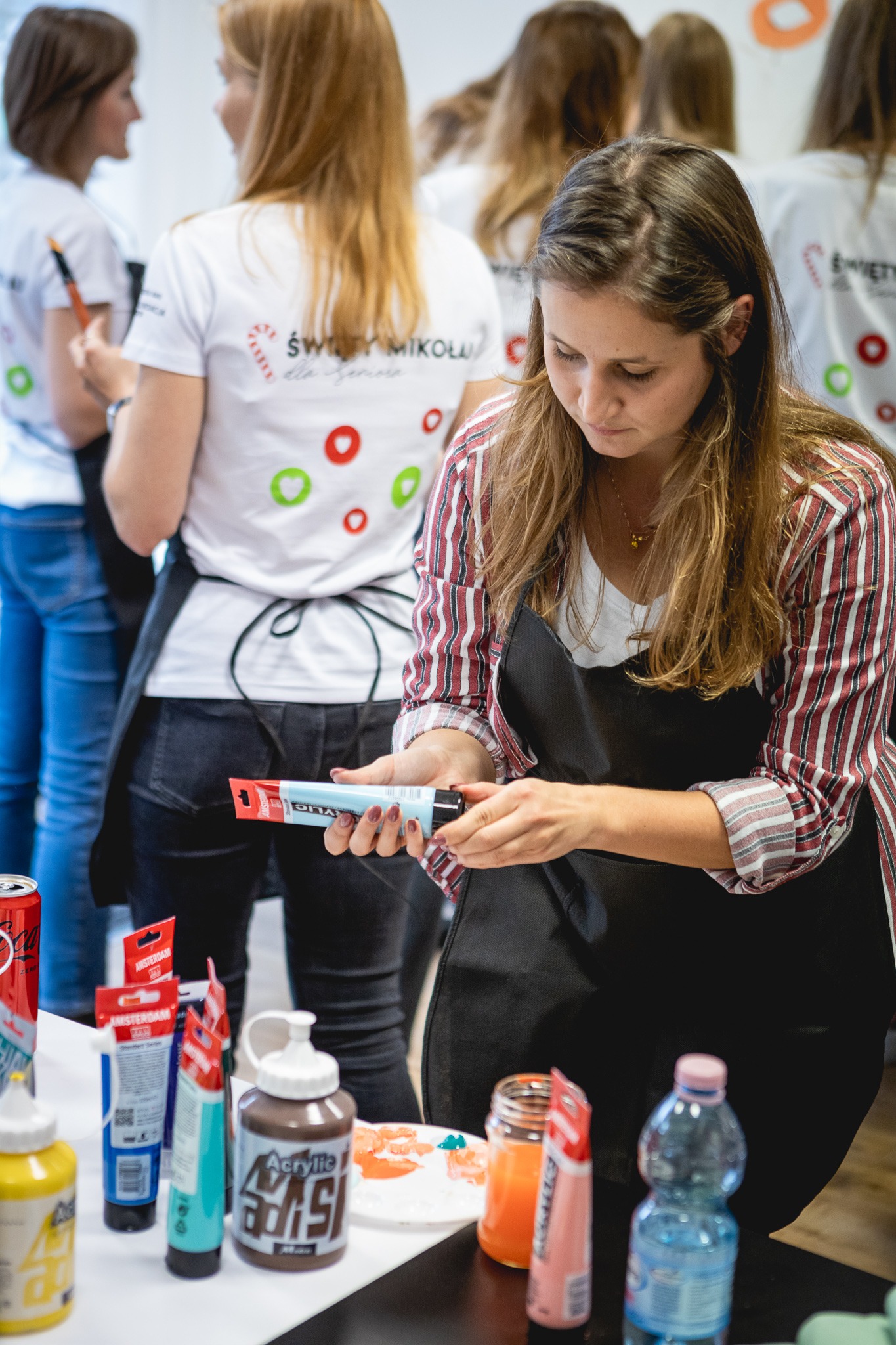 Kobieta w fartuchu przygotowuje farby akrylowe podczas warsztatów artystycznych, w tle grupa osób w koszulkach z logo Świąt Mikołajowych.
