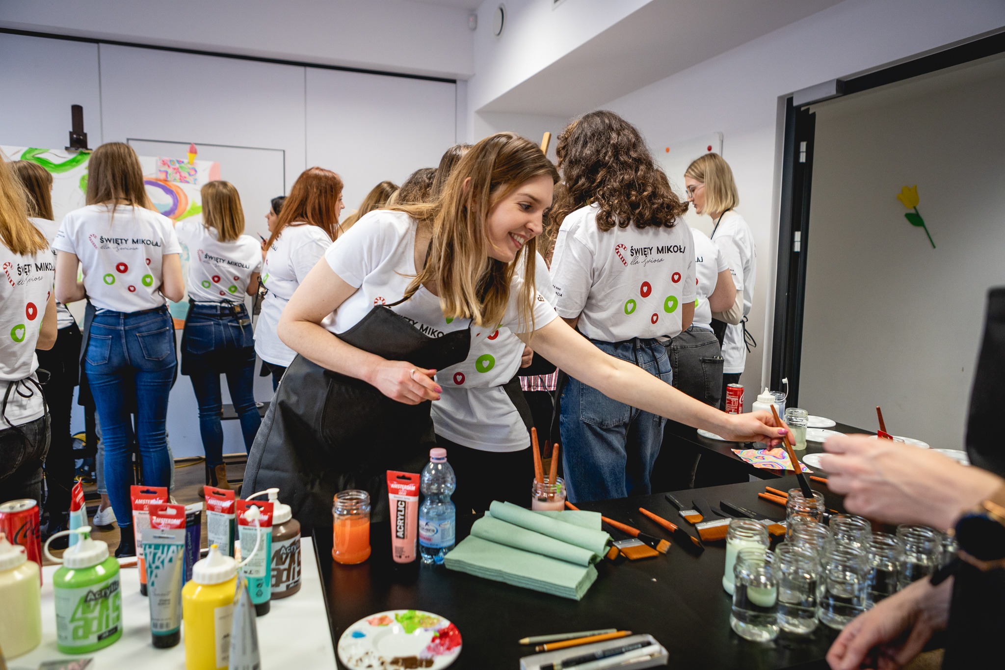 Grupa uśmiechniętych kobiet w białych koszulkach z logo 'Święty Mikołaj dla Seniora' bierze udział w warsztatach malarskich, używając pędzli i farb akrylowych na czarnych stołach.