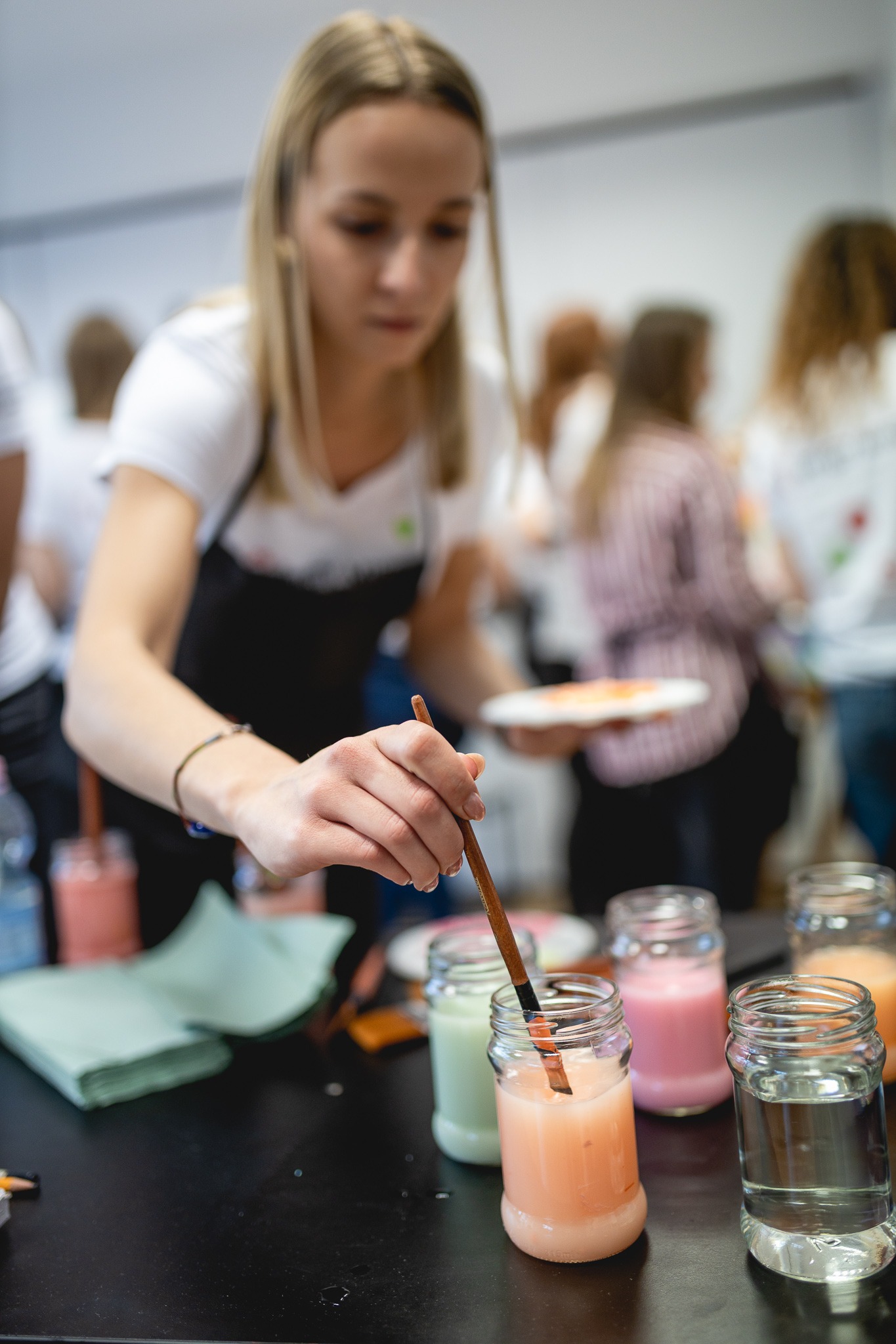 Młoda kobieta w czarnym fartuchu zanurza pędzel w słoiku z pomarańczową farbą podczas warsztatów malarskich dla grupy osób.