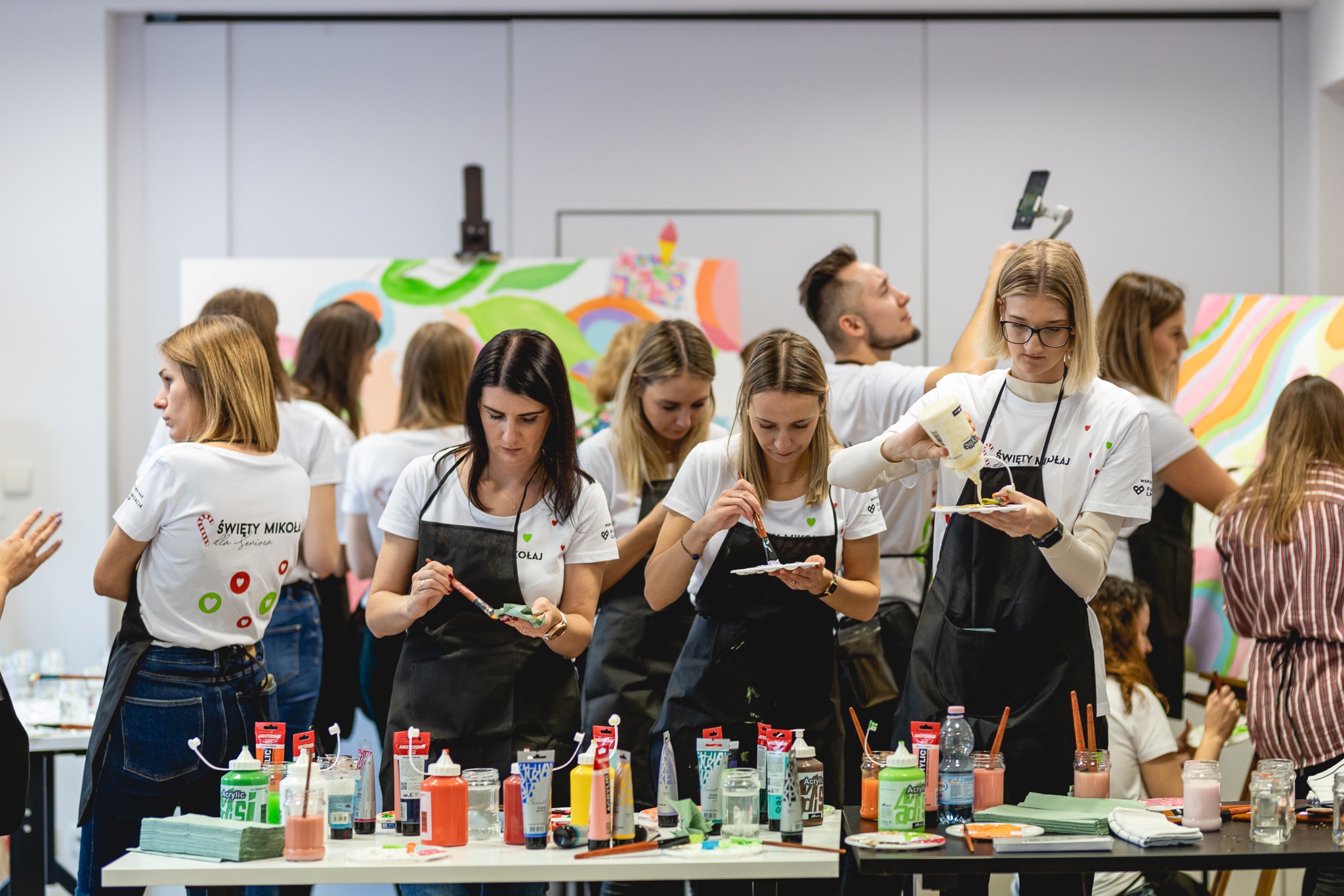 Grupa osób w białych koszulkach z napisem 'Święty Mikołaj', ubranych w czarne fartuchy, maluje obrazy podczas warsztatów artystycznych. Na stołach farby, pędzle i inne akcesoria malarskie.