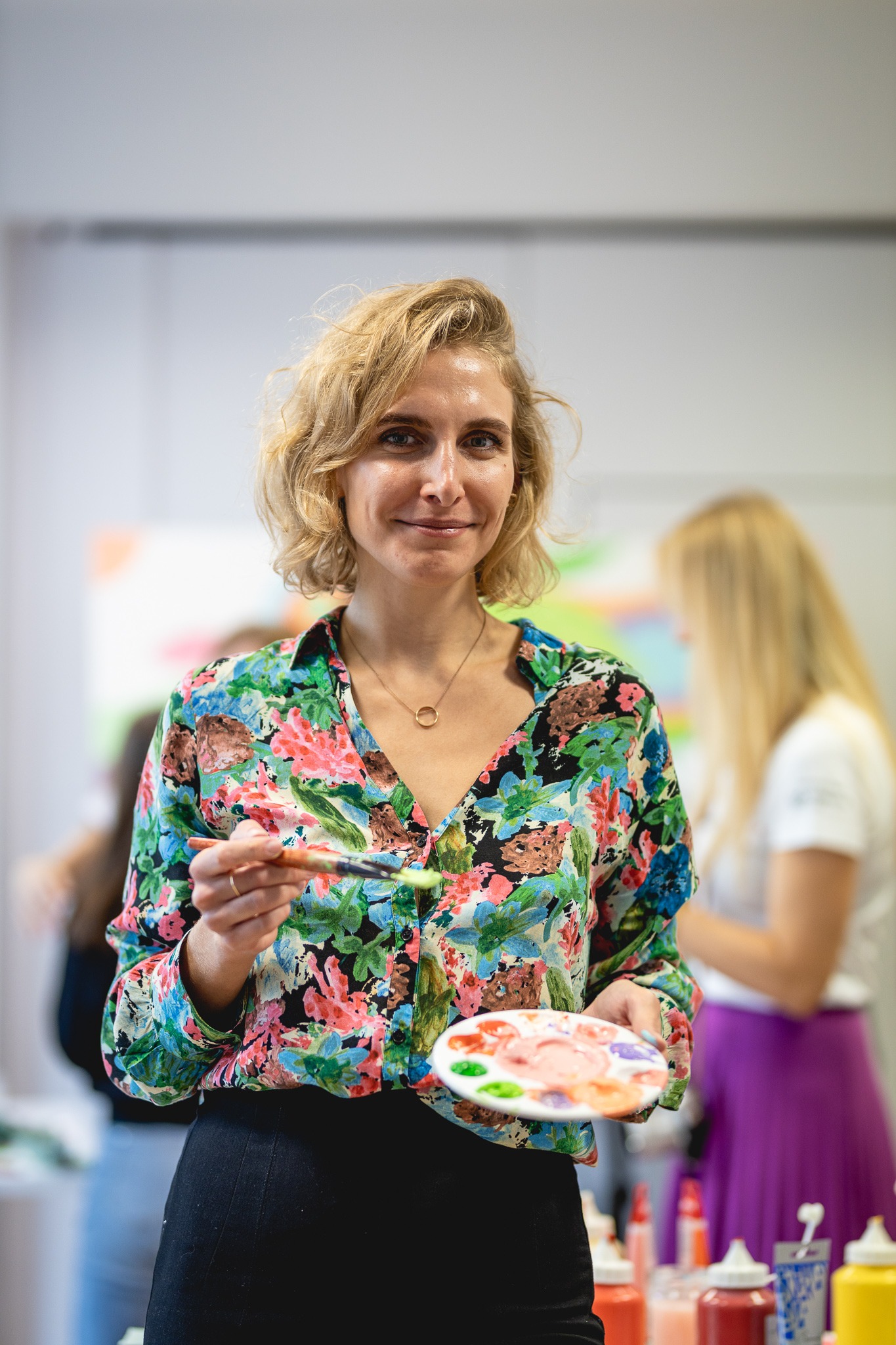 Uśmiechnięta blondynka w kwiecistej koszuli trzyma pędzel z zieloną farbą i paletę z farbami, w tle rozmazane postacie i butelki z farbami.
