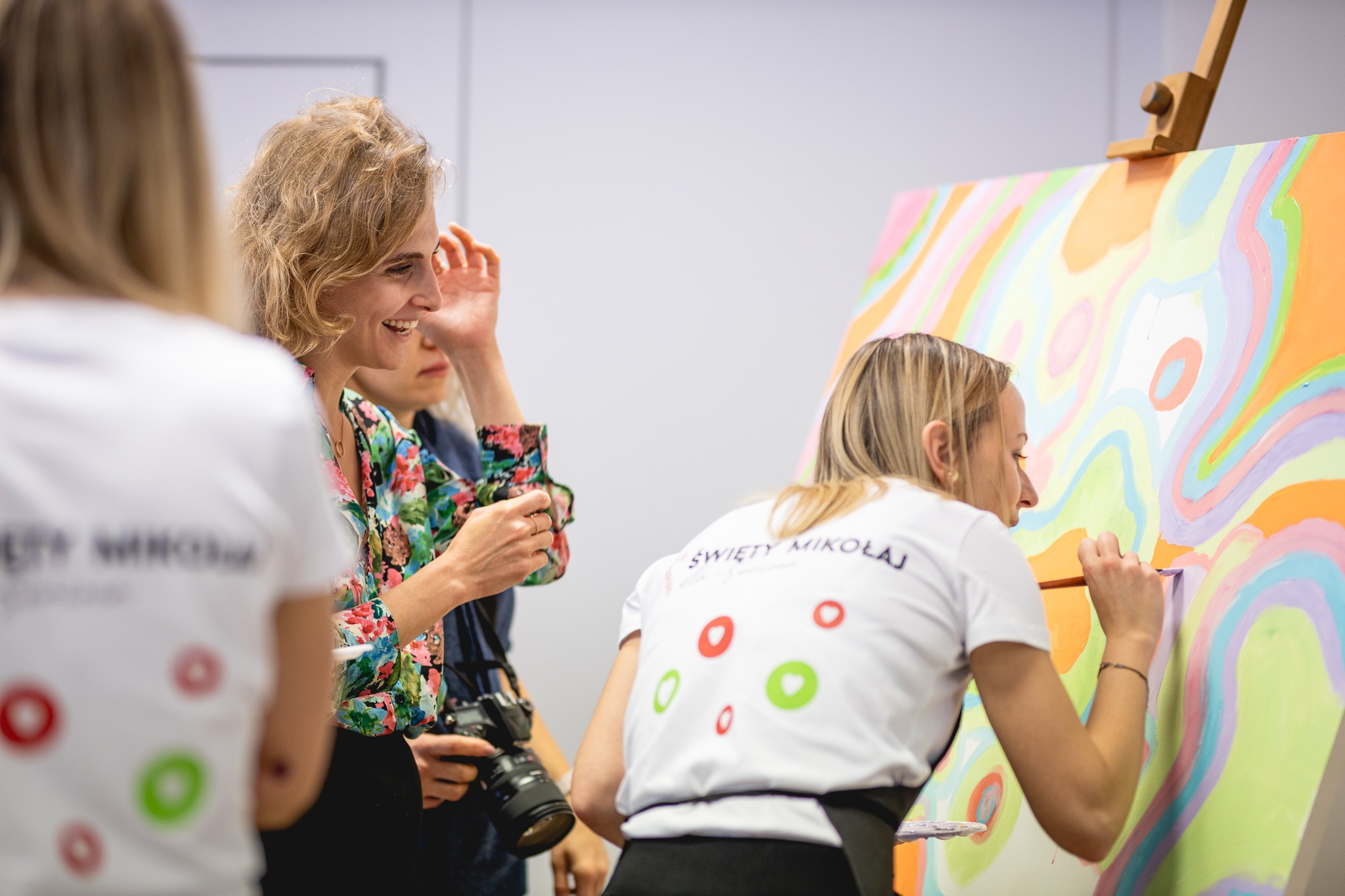 Warszawa: Kobieta maluje abstrakcyjny obraz na płótnie podczas warsztatów artystycznych, w tle uśmiechnięta kobieta z aparatem fotograficznym.
