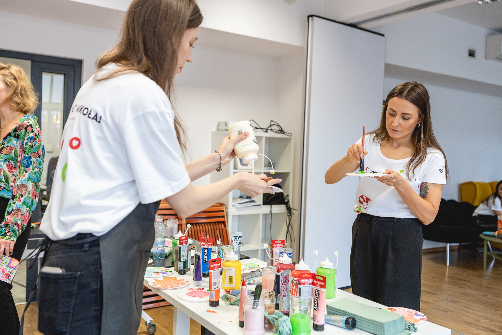Dwie kobiety przygotowują farby akrylowe na paletach. Jedna wyciska farbę z tubki, druga miesza farbę pędzlem na talerzyku. Na stole rozstawione różnokolorowe farby akrylowe w tubach i słoikach.