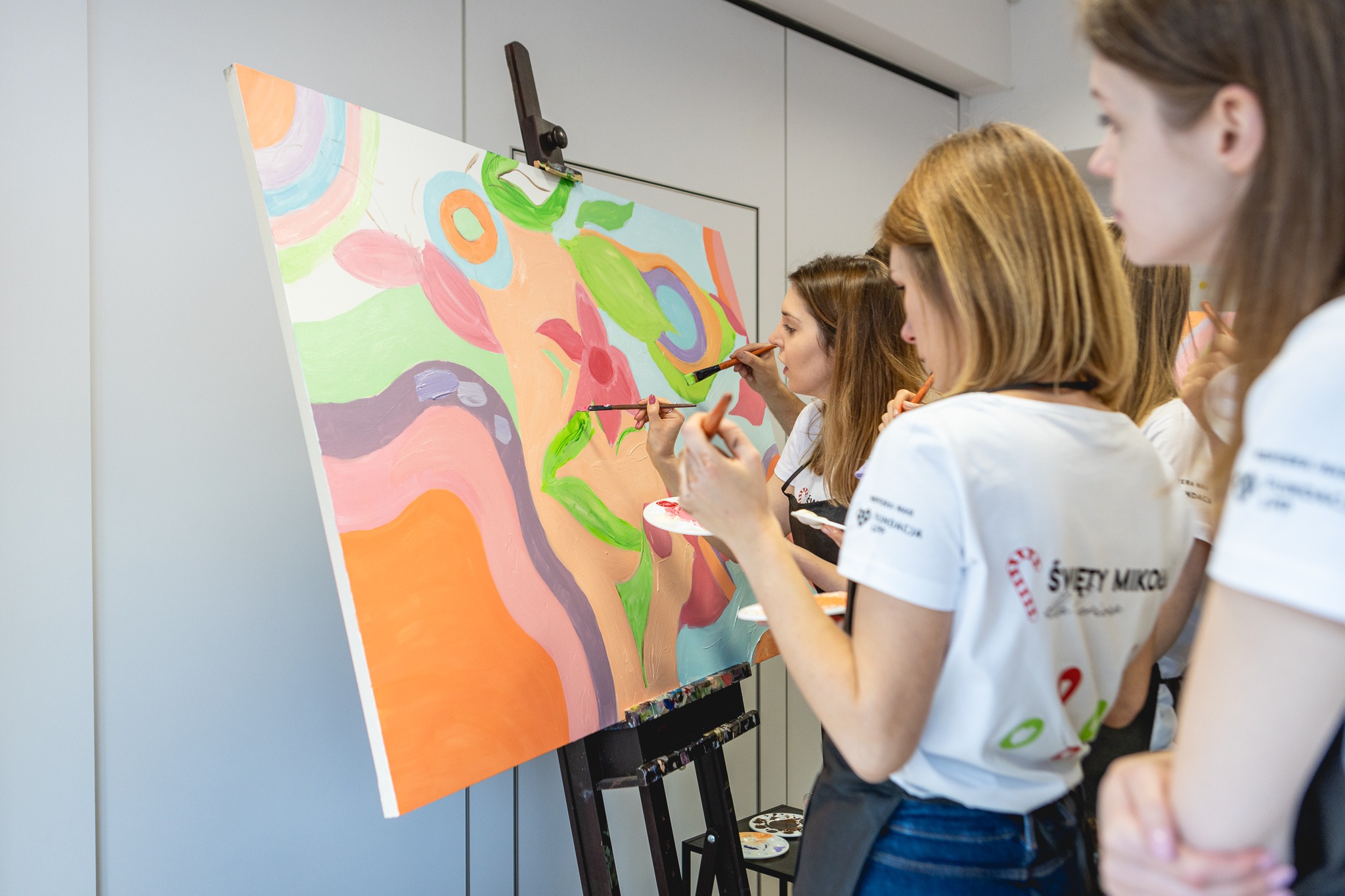 Grupa kobiet maluje abstrakcyjny obraz na sztaludze podczas warsztatów artystycznych, widoczne pędzle i palety z farbami, uczestniczki w identycznych koszulkach z logo.