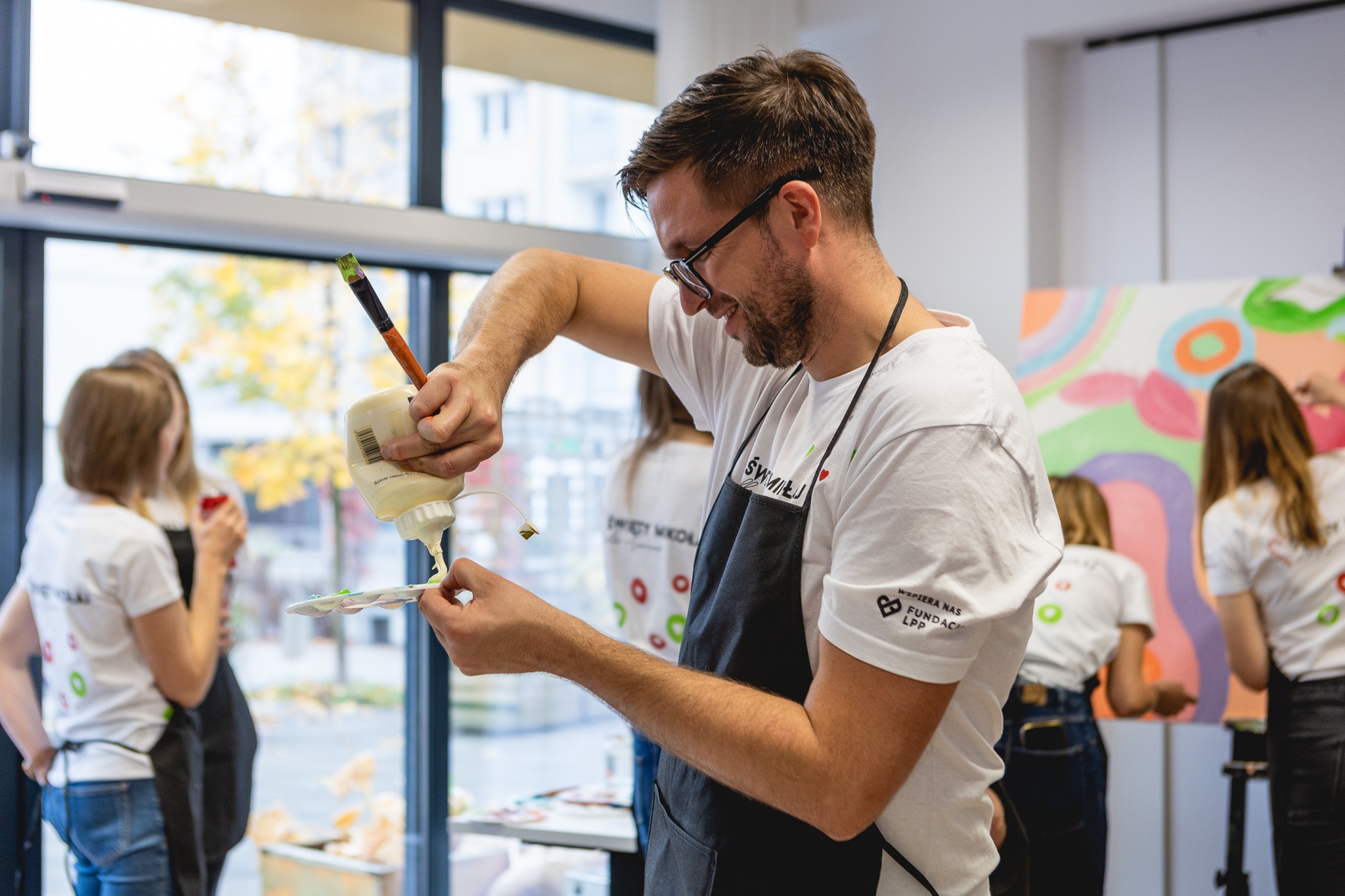 Mężczyzna w okularach i fartuchu nakłada farbę z tubki na paletę podczas warsztatów malarskich, w tle okno i inne osoby malujące obrazy.