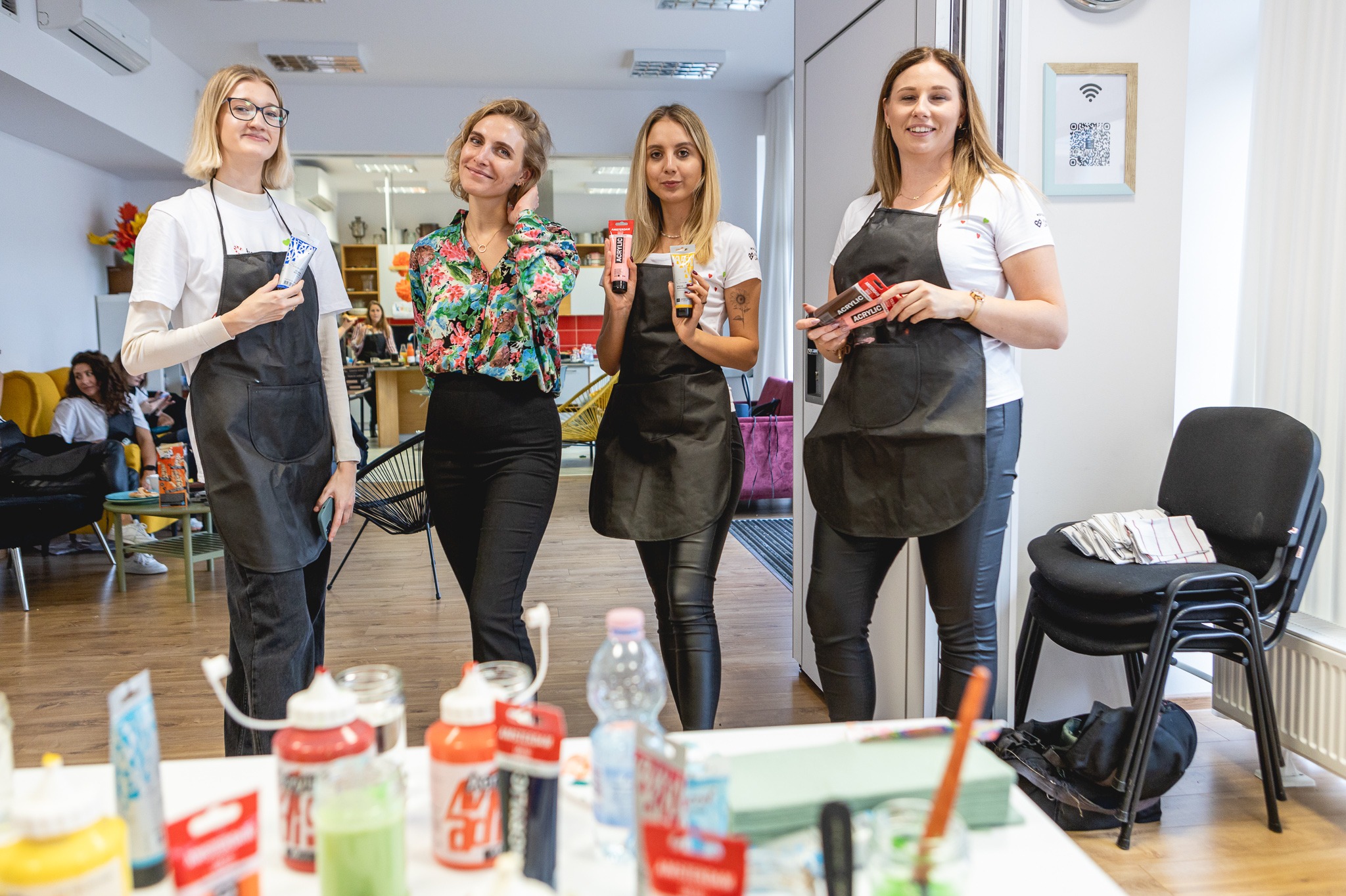 Cztery kobiety w fartuchach prezentują farby akrylowe w pracowni artystycznej, z widocznymi słoikami z farbami i innymi materiałami plastycznymi na stole, w tle siedzą ludzie.