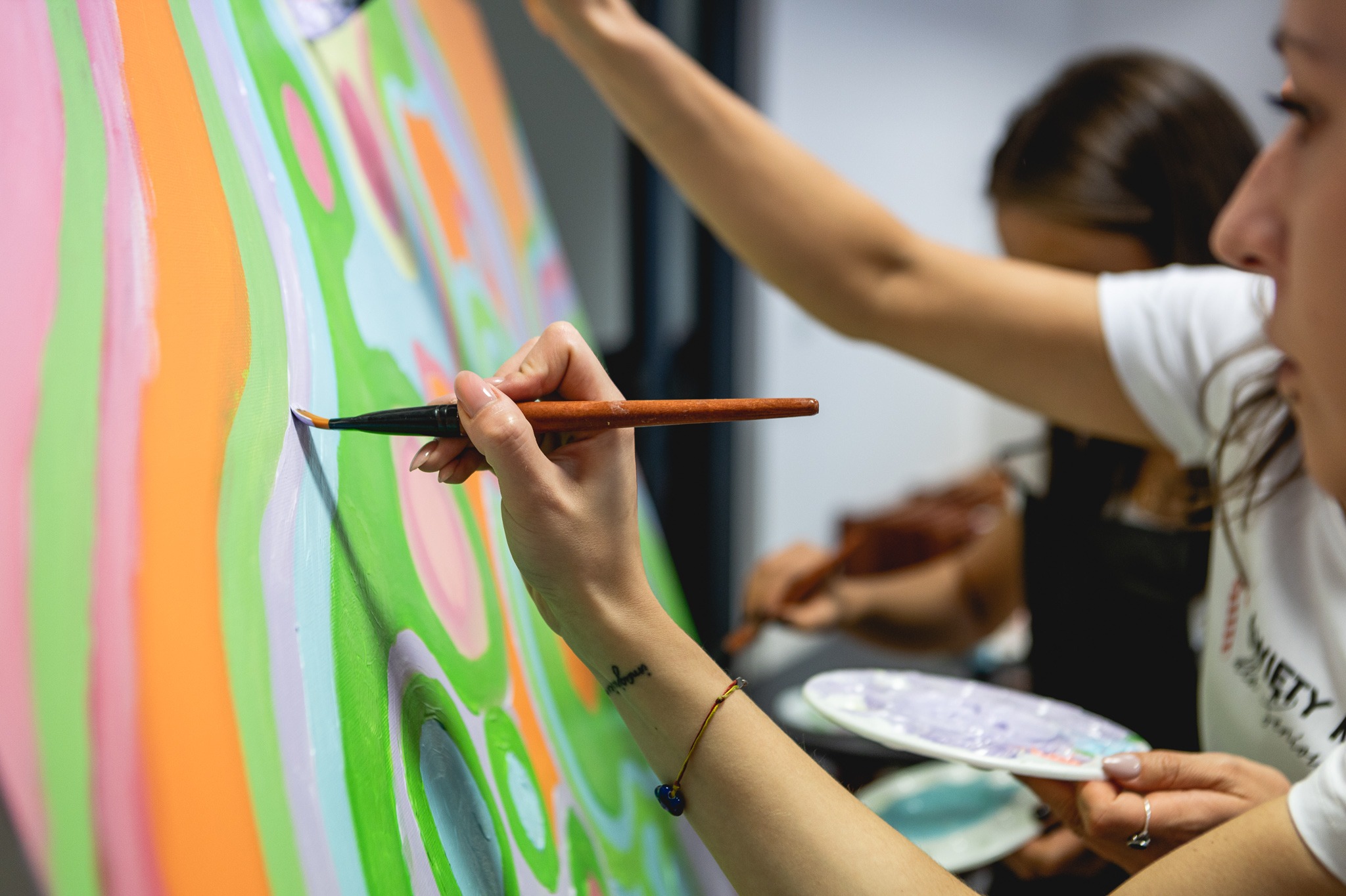 Dwie osoby malują abstrakcyjny obraz na płótnie, widoczny pędzel z farbą i paleta barw, ujęcie z bliska na dłoń z tatuażem.