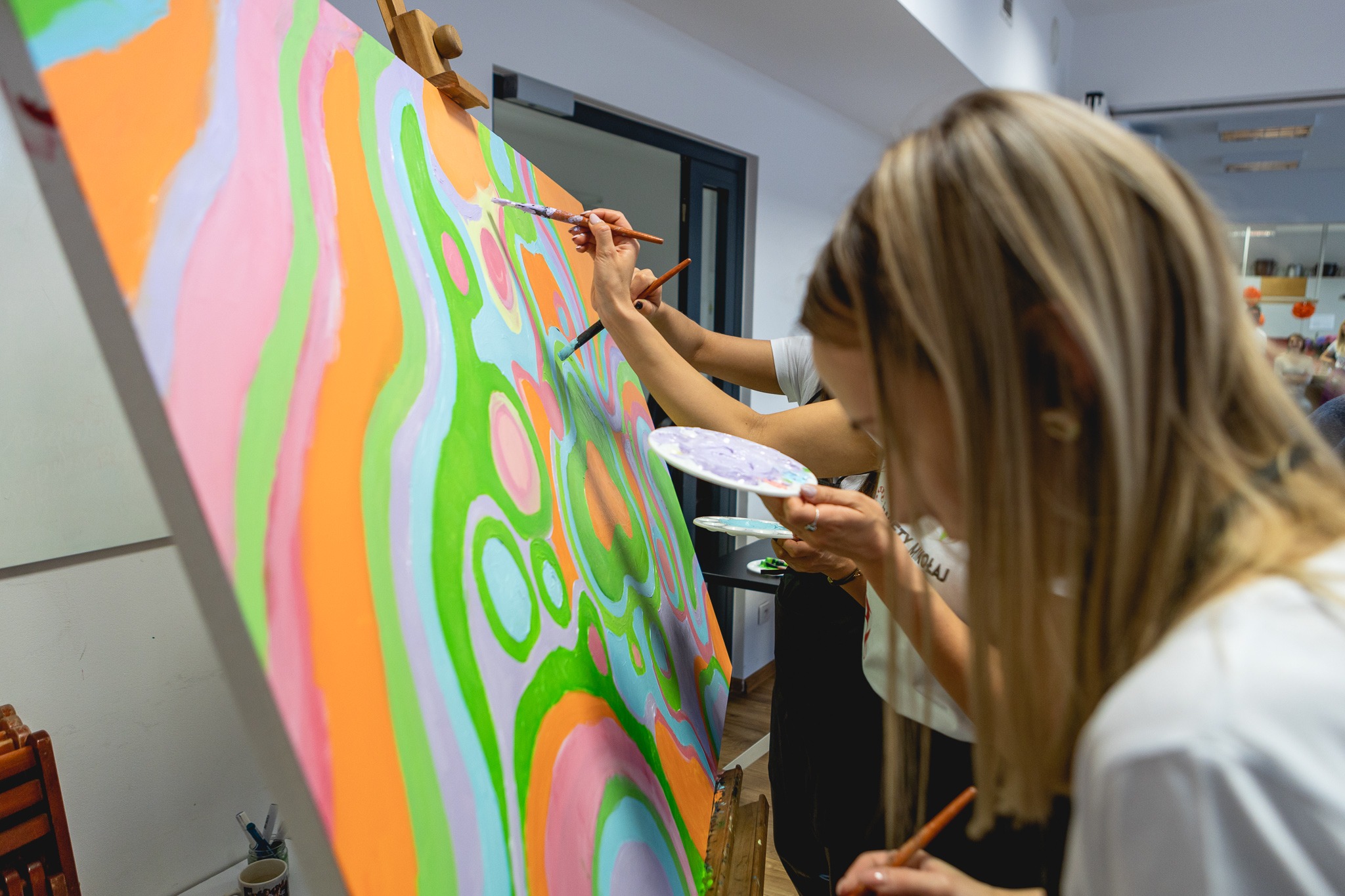 Grupa osób wspólnie maluje abstrakcyjny obraz na płótnie, używając pędzli i palet z farbami podczas warsztatów artystycznych.