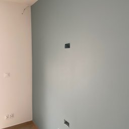 Patryk Kaśków - Świeżo pomalowana ściana w odcieniach szarości z widocznymi puszkami elektrycznymi i wystającym kablem w rogu, podłoga wykończona panelami w jodełkę.