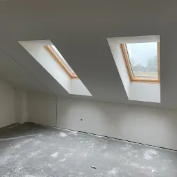 Poddasze w trakcie prac wykończeniowych: szpachlowane ściany i sufit z dwoma oknami dachowymi, widoczny laser krzyżowy na betonowej podłodze.