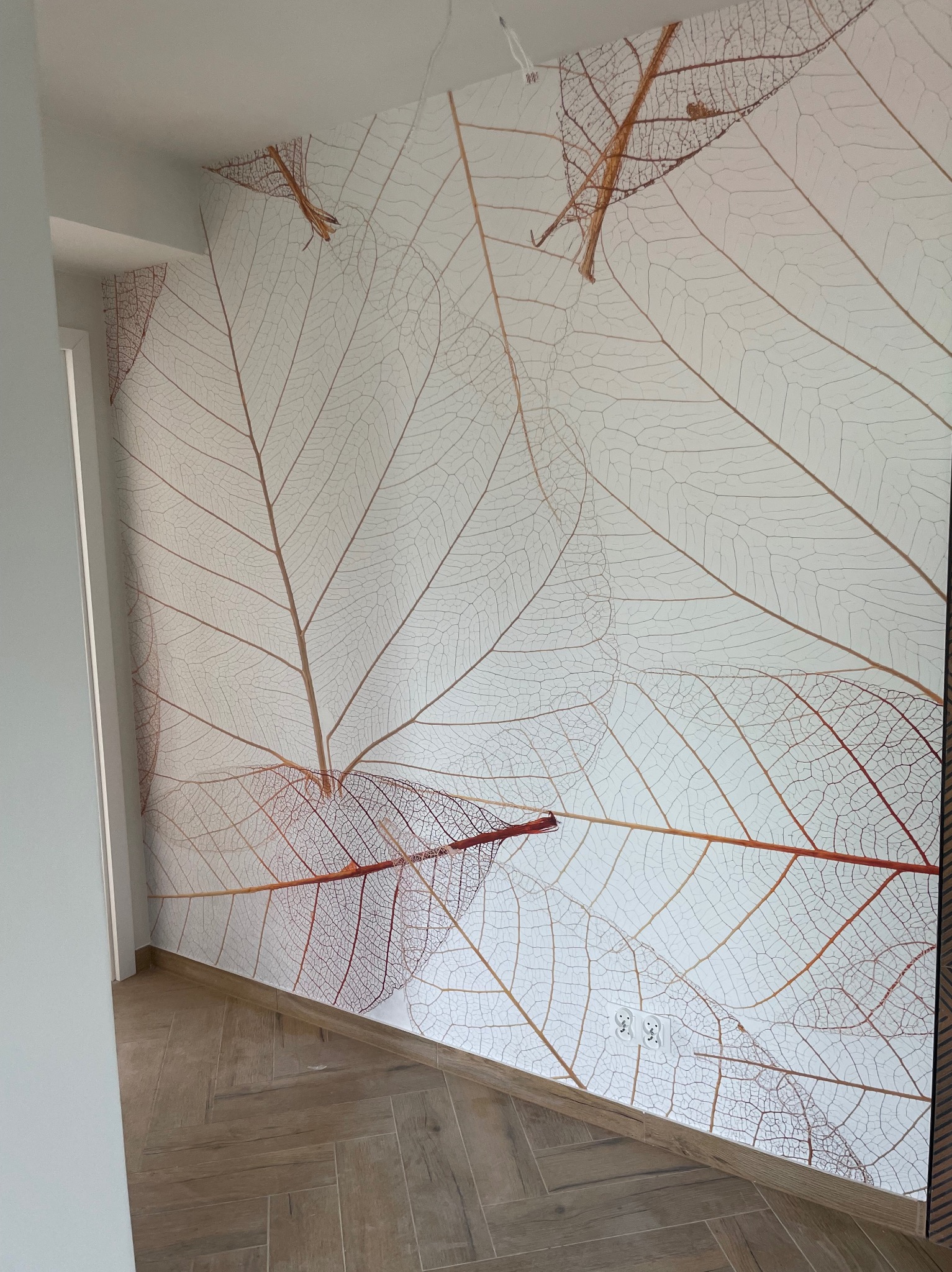 Ściana w korytarzu pokryta tapetą z motywem szkieletów liści w odcieniach brązu i beżu, ułożona nad podłogą z płytek imitujących drewno, widoczne gniazdka elektryczne.