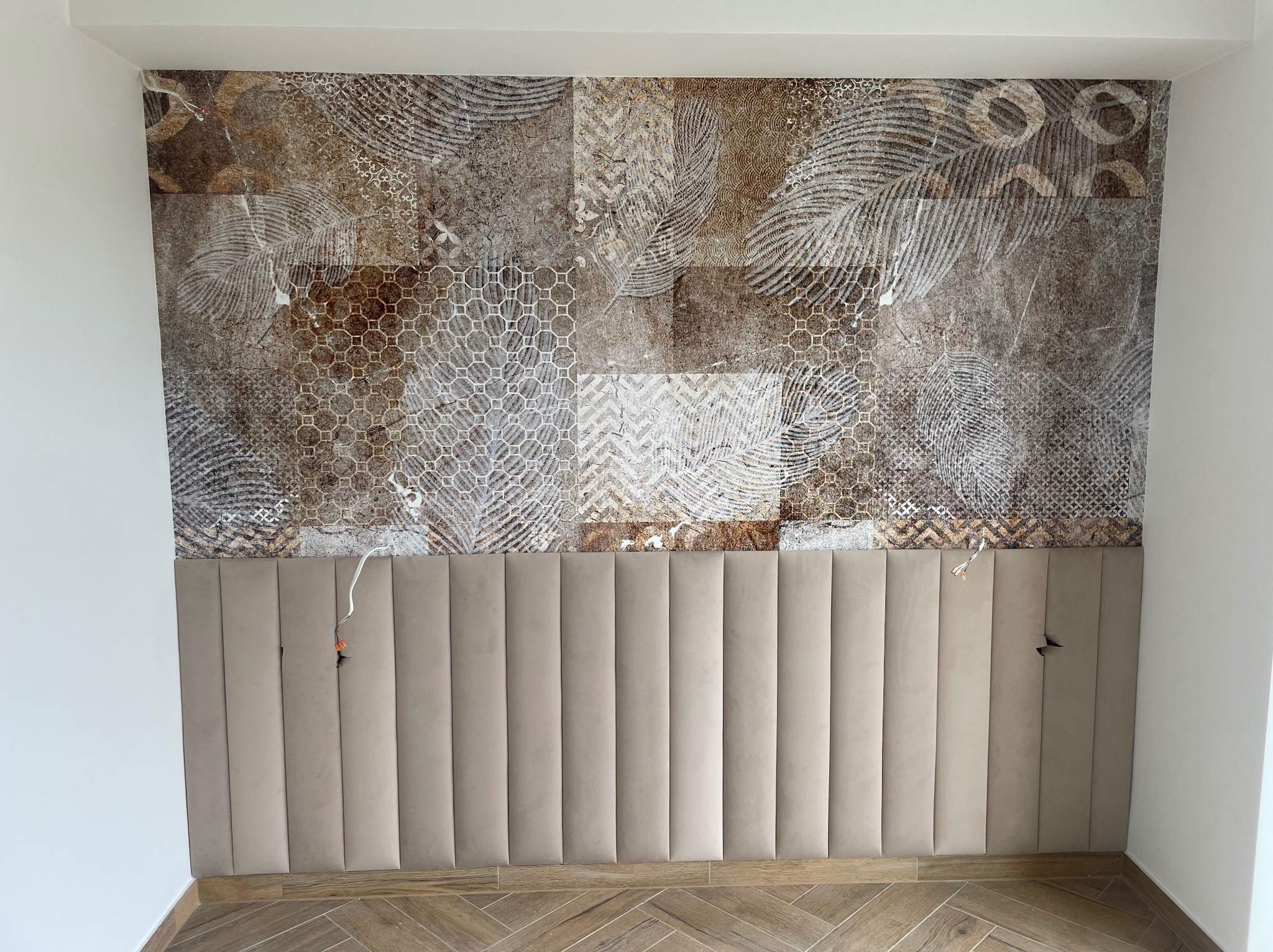 Panel tapicerowany w kolorze beżowym, zamontowany na ścianie z dekoracyjną brązowo-beżową tapetą z motywami geometrycznymi i roślinnymi, widoczne wystające kable elektryczne.