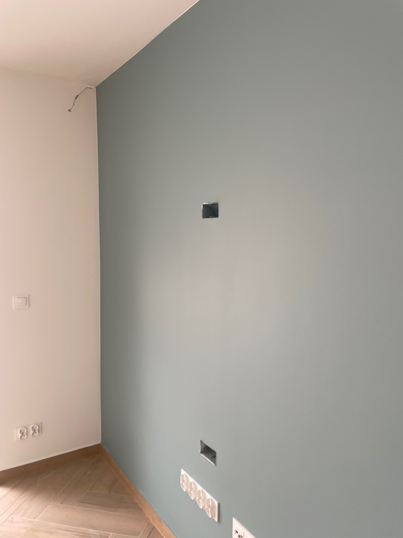 Świeżo pomalowana ściana w odcieniach szarości z widocznymi puszkami elektrycznymi i wystającym kablem w rogu, podłoga wykończona panelami w jodełkę.