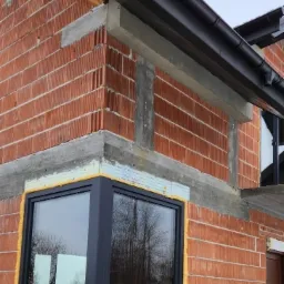 Montaż narożnego okna w surowym murze z czerwonej cegły, widoczna piana montażowa i betonowe nadproże, odbicie drzew w szybie.