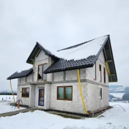 Dom w stanie surowym zamkniętym w Łososinie Dolnej, z widocznymi brązowymi ramami okiennymi i niebieskimi drzwiami wejściowymi, pokryty śniegiem na dachu i otoczony zimowym krajobrazem.