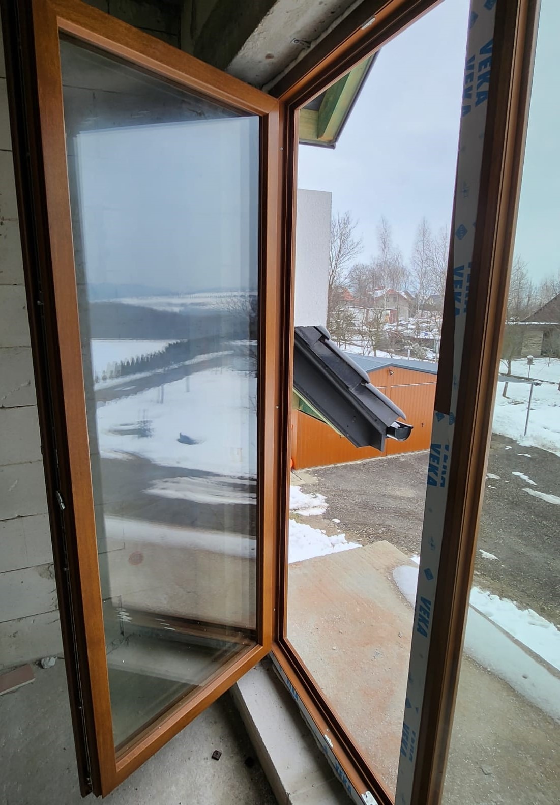 Otwarty drewnopodobny profil okienny z widokiem na zimowy krajobraz i fragment dachu sąsiedniego budynku, widoczna folia ochronna na ramie.