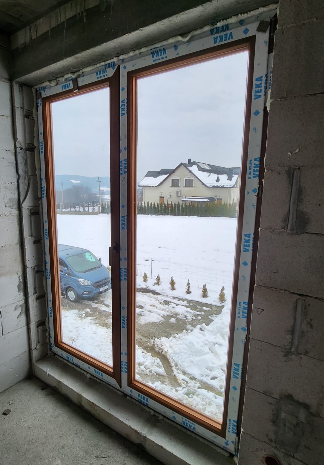 Montaż okna z drewnopodobną ramą w surowym stanie, widok na zasypany śniegiem krajobraz z domem i niebieskim busem na podjeździe.