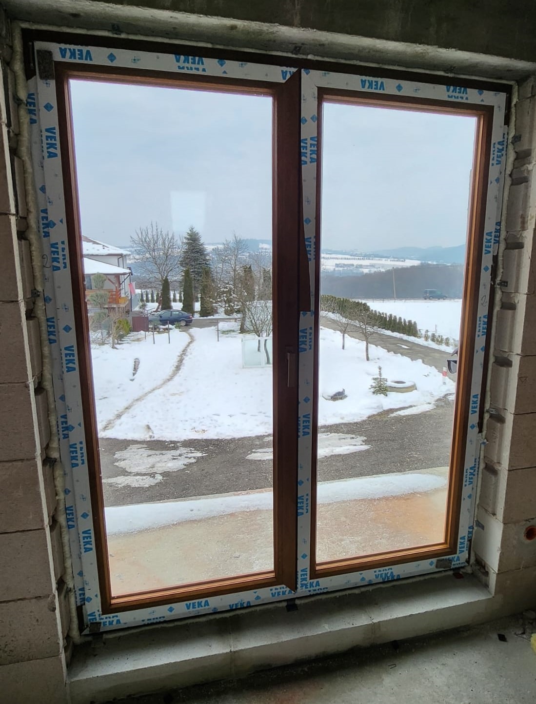Brązowe dwuskrzydłowe okno z widokiem na zimowy krajobraz z ośnieżonymi drzewami i drogą, zamontowane w surowym murze z widoczną pianką montażową i folią ochronną z logo VEKA.