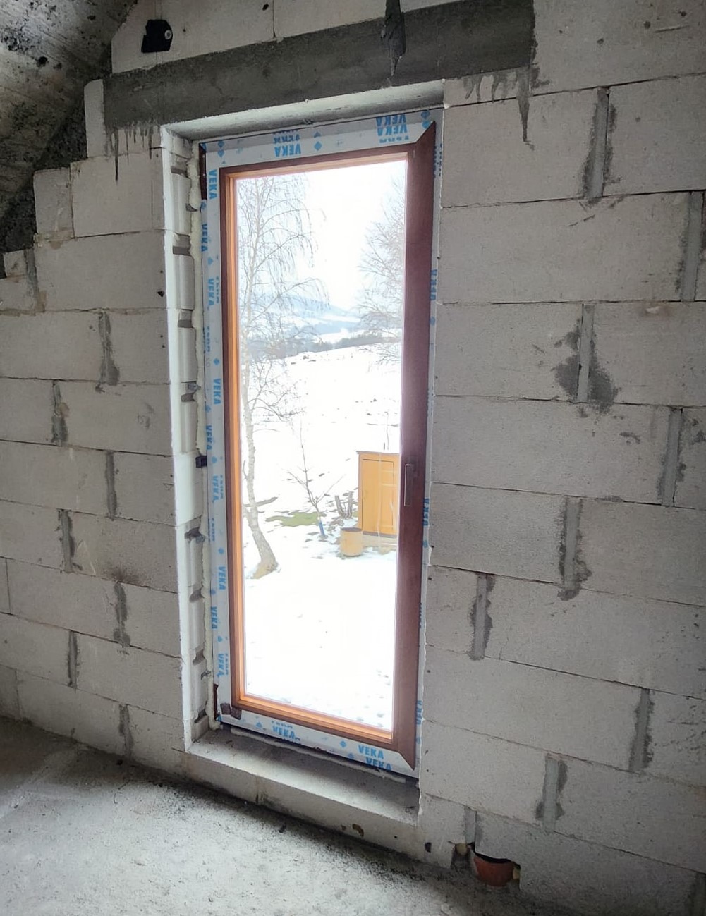 Montaż okna w surowym stanie, widoczne białe bloczki konstrukcyjne i zimowy krajobraz za oknem.