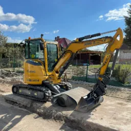 Żółta mini koparka XCMG XE35E na placu budowy, widoczne wykopane koryto, rury drenarskie i częściowo wykończony budynek w tle.