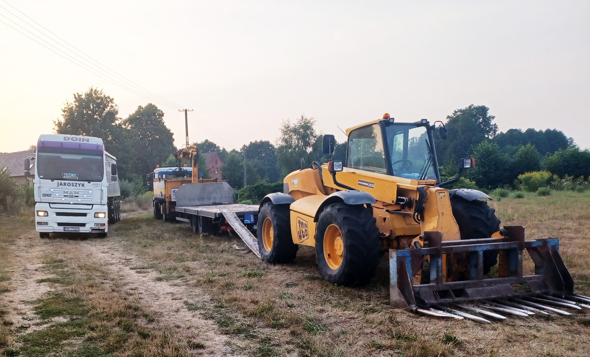 Żółta ładowarka teleskopowa JCB z widłami na nierównym terenie, transportowana na niskopodwoziowej przyczepie za ciężarówką MAN firmy Jaroszyk, widok z boku w pochmurny dzień.