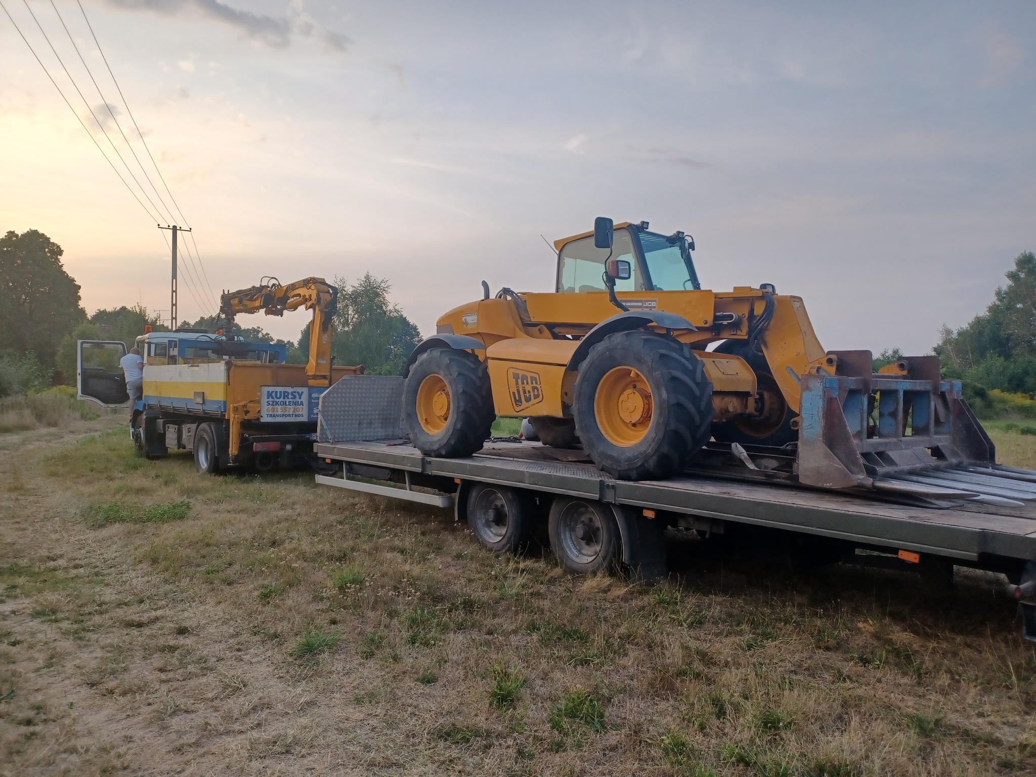 Żółta ładowarka teleskopowa JCB przewożona na niskopodwoziowej przyczepie przez ciężarówkę z dźwigiem HDS, na tle łąki i zachodzącego słońca.