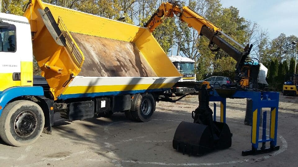 Żółto-biało-niebieska wywrotka z zamontowanym ramieniem dźwigu i łyżką koparki na placu budowy, w tle inne pojazdy i drzewa.