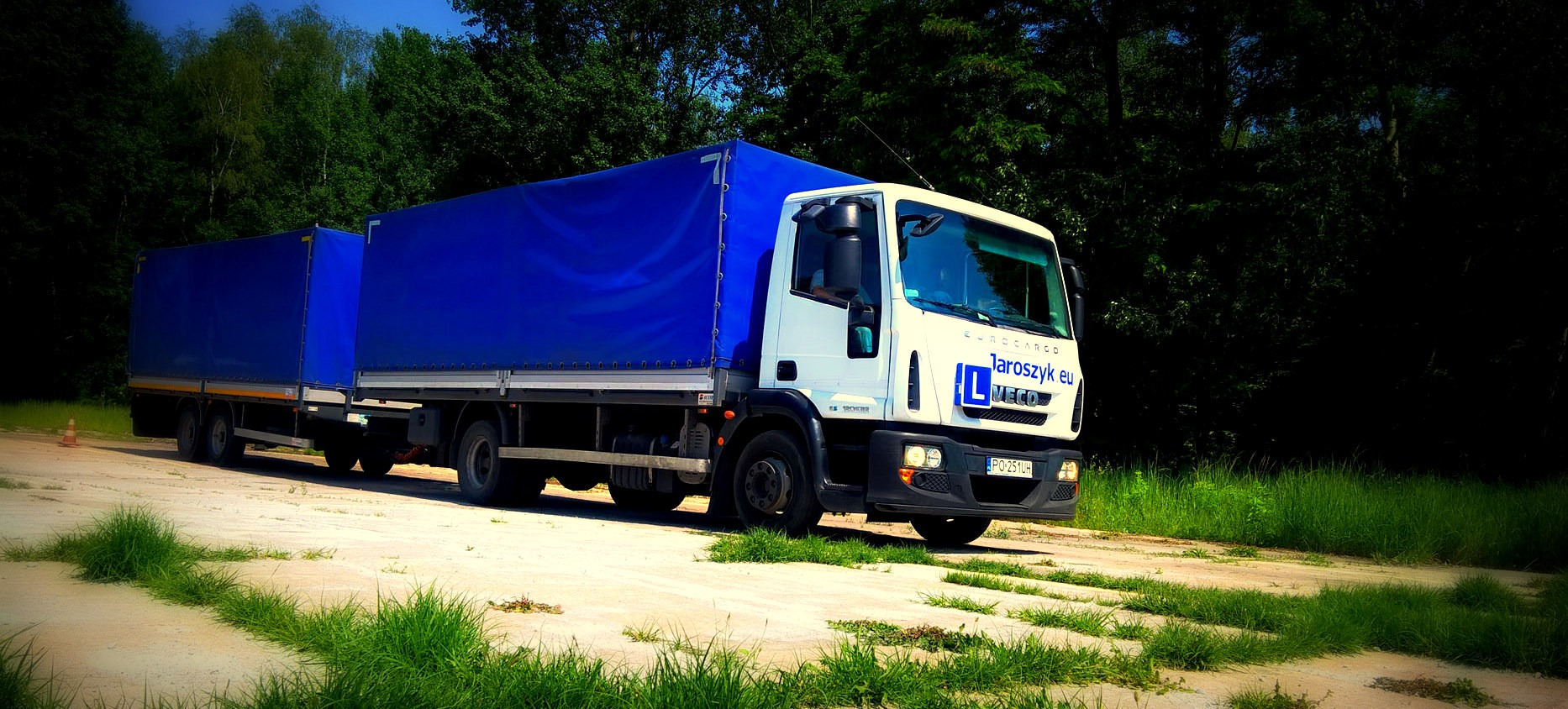 Biała ciężarówka z niebieską plandeką na naczepie, zaparkowana na ubitej drodze gruntowej z zieloną trawą, w tle ciemny las.