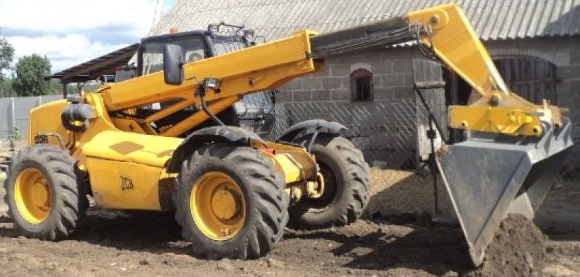 Żółta ładowarka teleskopowa JCB z łyżką nabierająca ziemię na placu budowy, widoczny budynek gospodarczy w tle.