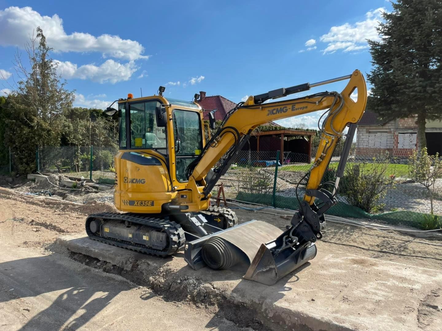 Żółta mini koparka XCMG XE35E na placu budowy, widoczne wykopane koryto, rury drenarskie i częściowo wykończony budynek w tle.