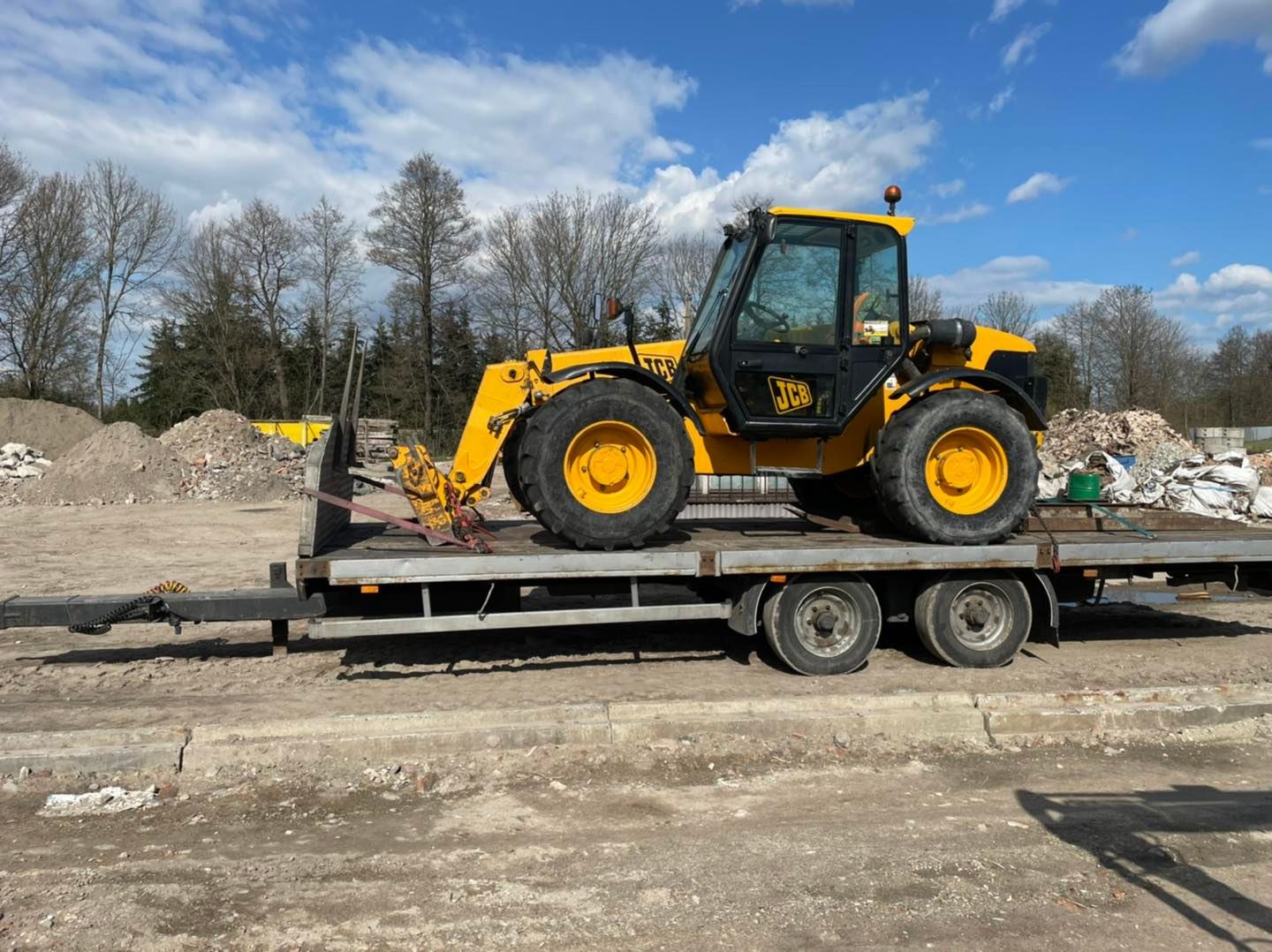 Żółta ładowarka teleskopowa JCB zabezpieczona pasami na niskopodwoziowej przyczepie, w tle sterta gruzu i drzewa bez liści, błękitne niebo z chmurami.