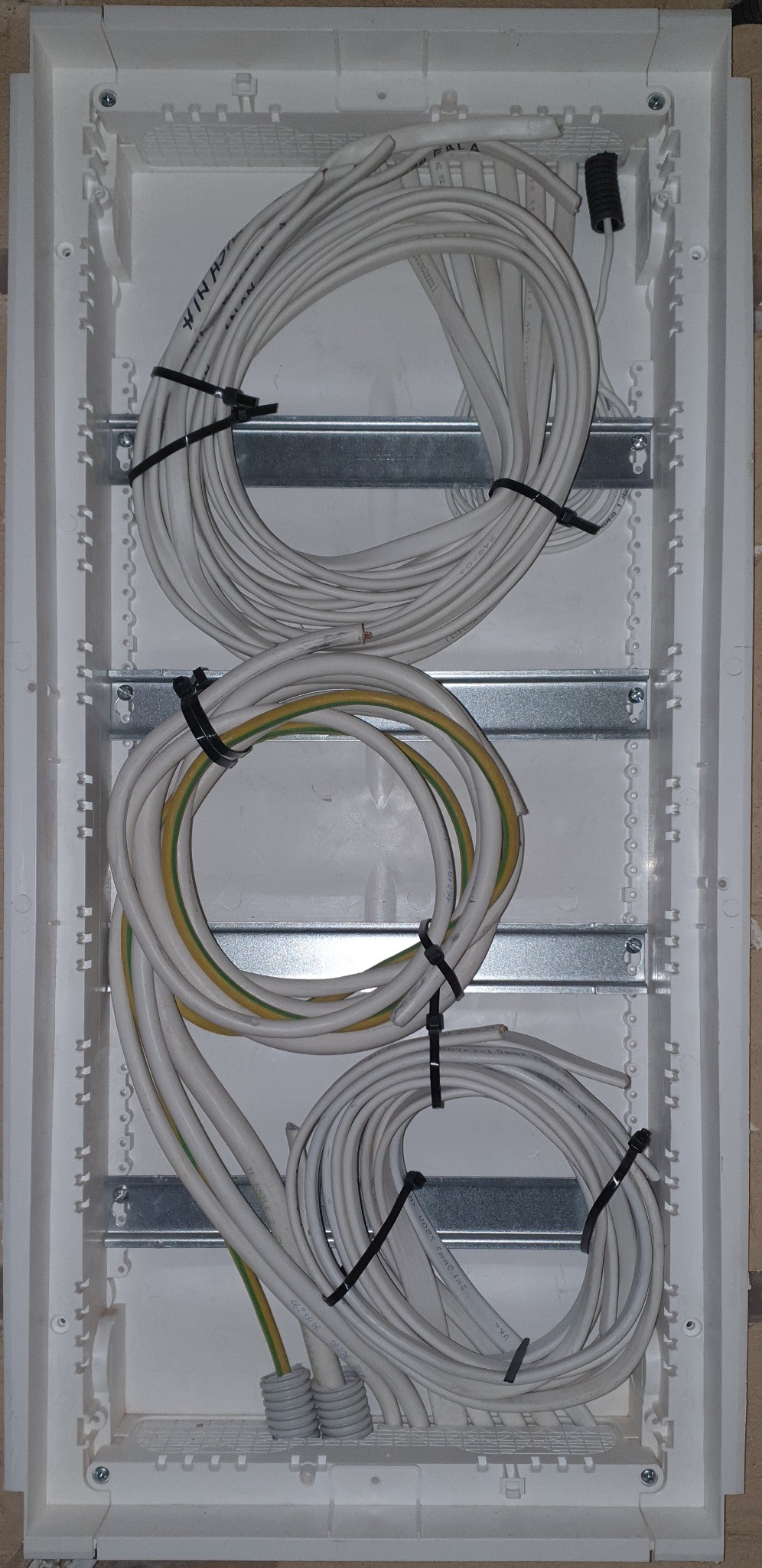 Rozdzielnica elektryczna w trakcie montażu, widoczne kable ułożone w pętle i przymocowane opaskami zaciskowymi do metalowych szyn wewnątrz obudowy z tworzywa.