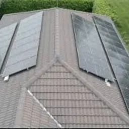 Widok z góry na dach domu z trzema rzędami paneli fotowoltaicznych o różnym kącie nachylenia, otoczony zielenią drzew i trawnika.