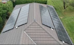 Widok z góry na dach domu z trzema rzędami paneli fotowoltaicznych o różnym kącie nachylenia, otoczony zielenią drzew i trawnika.