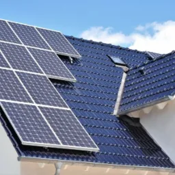 Instalacja paneli słonecznych na dachu domu jednorodzinnego z ciemną dachówką, widoczne okno dachowe i logo firmy na ścianie budynku.