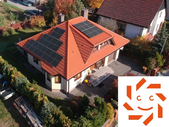 Dom jednorodzinny z czerwoną dachówką, na której zamontowano panele fotowoltaiczne; widok z góry, obok widoczne logo firmy.