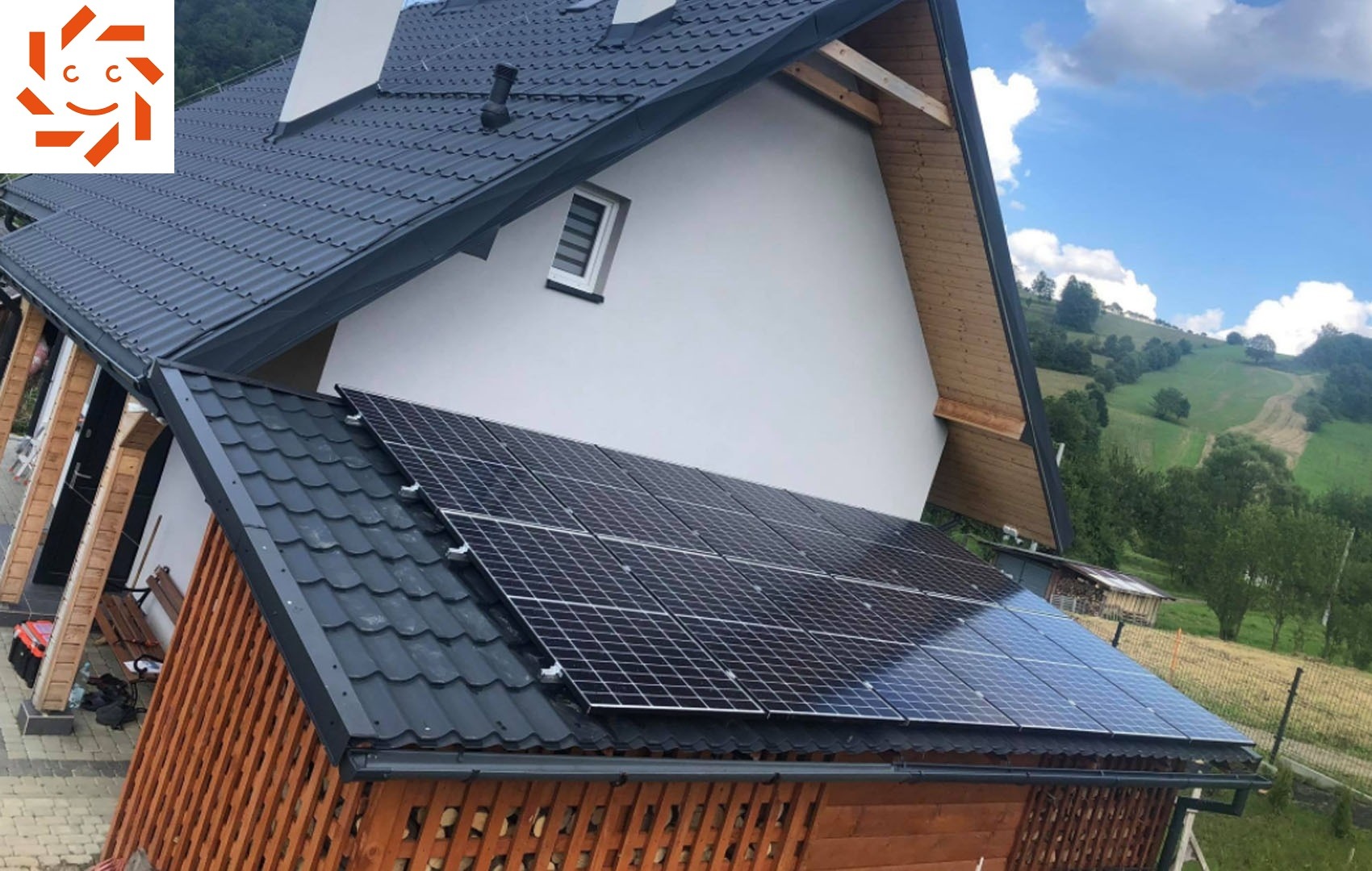 Panele słoneczne na dachu domu z drewnianą elewacją, w tle zielone wzgórza i błękitne niebo z chmurami.