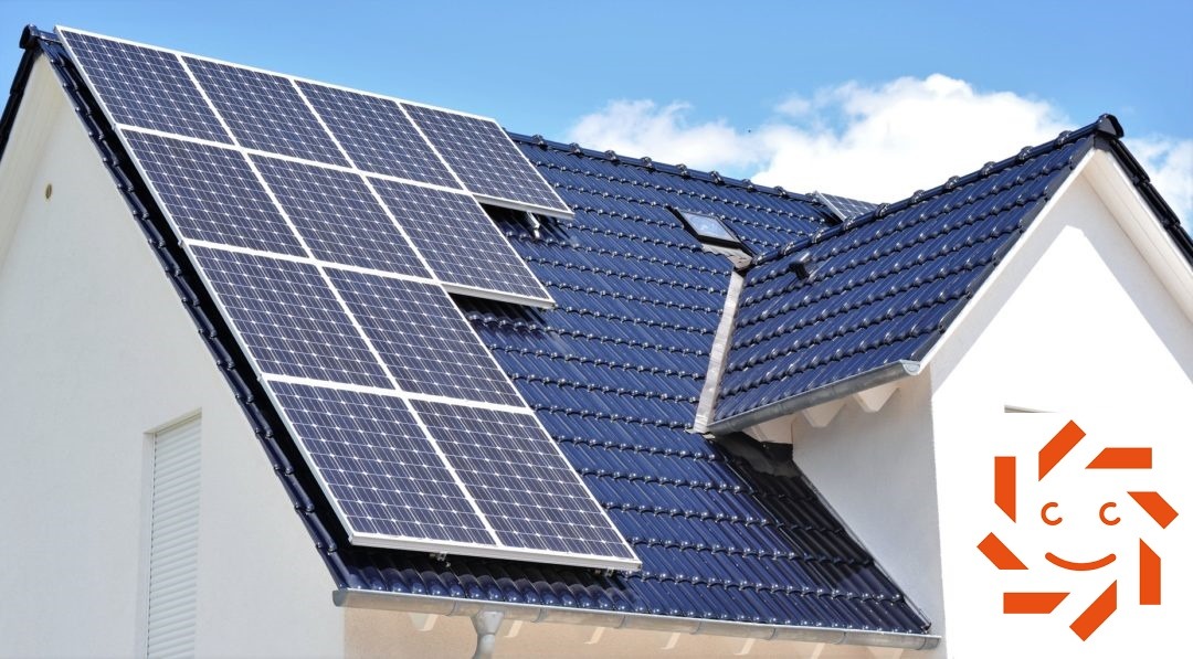 Instalacja paneli słonecznych na dachu domu jednorodzinnego z ciemną dachówką, widoczne okno dachowe i logo firmy na ścianie budynku.