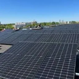 Instalacja paneli słonecznych na płaskim dachu budynku, widok z góry, w tle zabudowa miejska i błękitne niebo.