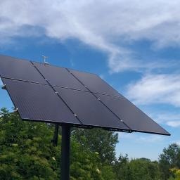 Panel słoneczny ustawiony pod kątem na tle błękitnego nieba z chmurami, widoczny z dołu, otoczony zielenią drzew.