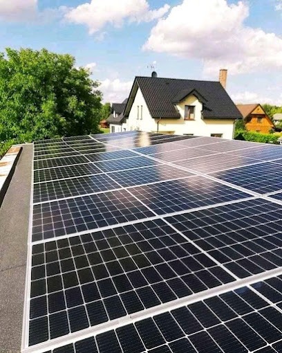 Panele fotowoltaiczne na płaskim dachu z widokiem na dom z ciemnym dachem i zielone drzewa w tle, odbicie nieba w panelach.