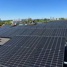 Instalacja paneli słonecznych na płaskim dachu budynku, widok z góry, w tle zabudowa miejska i błękitne niebo.