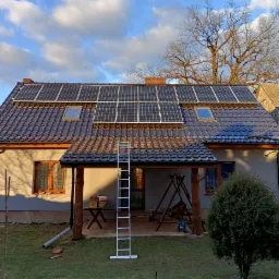 Panele słoneczne na brązowym dachu domu jednorodzinnego, z widoczną drabiną opartą o werandę i pochmurnym niebem w tle.
