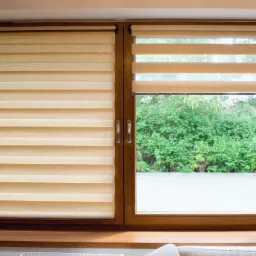 Drewniane okno z zamontowanymi beżowymi roletami z poziomymi pasami, częściowo zasłoniętymi, z widokiem na zieleń za oknem.