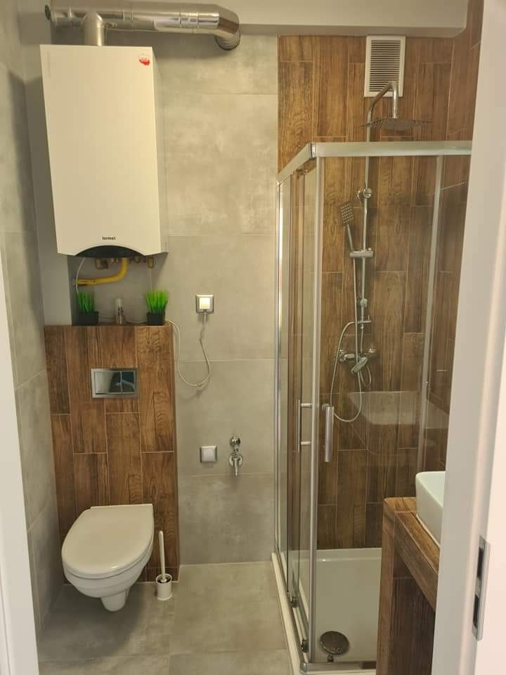 Nowoczesna łazienka z kabiną prysznicową, wiszącą toaletą i piecem gazowym, wykończona płytkami imitującymi drewno i szarym gresem.