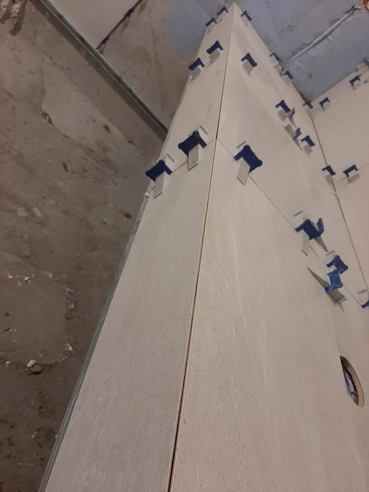 Montaż płytek ceramicznych na ścianie w rogu pomieszczenia, z użyciem niebieskich klipsów poziomujących, widoczne surowe ściany i niedokończony sufit.